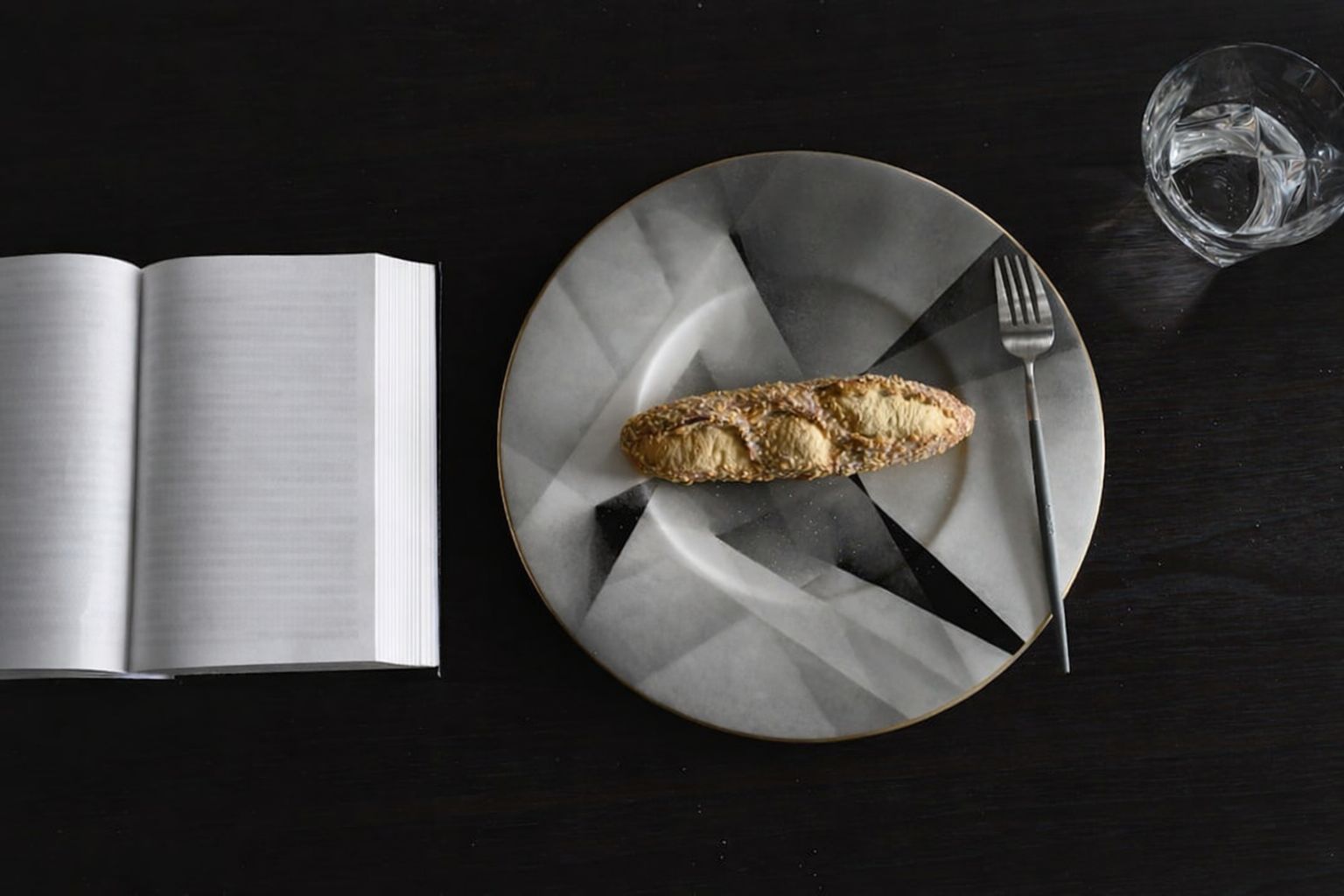 Bok, bröd på tallrik, gaffel och vattenglas på mörkt bord
