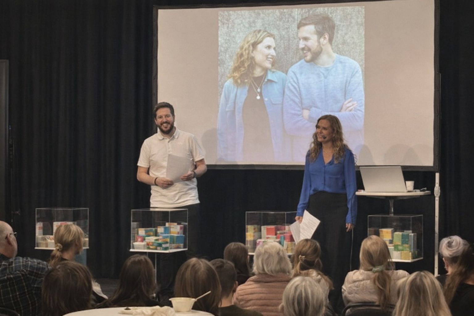 Två personer presenterar glutenfria matkassar för en publik framför en projektionsduk.